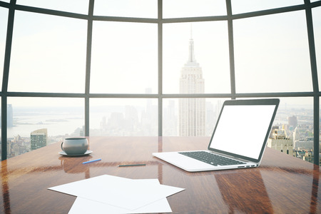 Closeup of shiny wooden desktop with blank white laptop, coffee cup and paper sheets with window and New York city view in the background. Mock up, 3D Renderingの写真素材