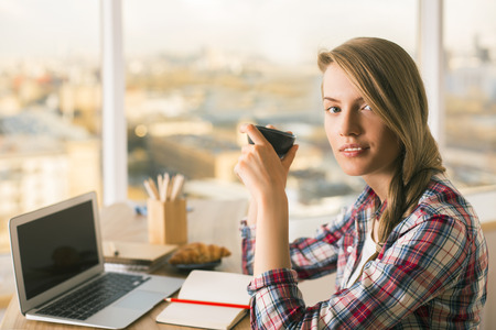 Pretty woman drinking coffee at office desk with laptop. Charming blonde woman having a break from working on laptop. Attractive girl doing homework on her laptop over cup of coffeeの写真素材