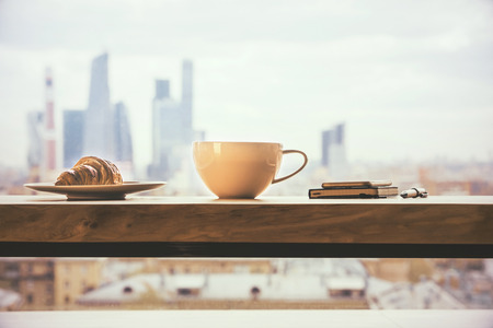 Closeup of round coffee mug, croissant and phone with diary on wooden desktop and city background