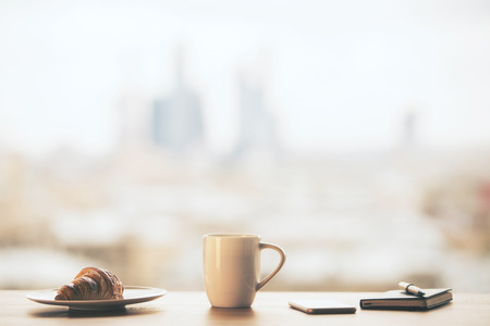 Table with croissant on plate, tall coffee cup, smartphone and notepad with pen on blurry city backgroundの写真素材