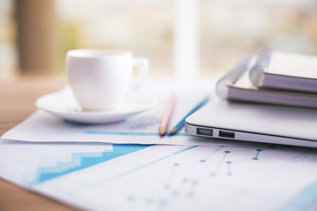 Closeup of coffee cup, business review, pencils, notepads and laptop on office desktopの写真素材