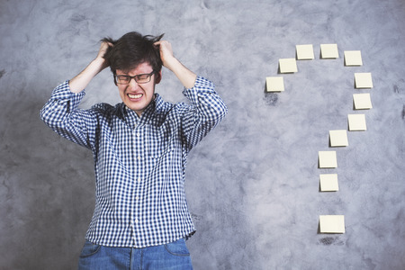 Stressed young man with hands on head standing next to sticker question mark glued onto concrete...
