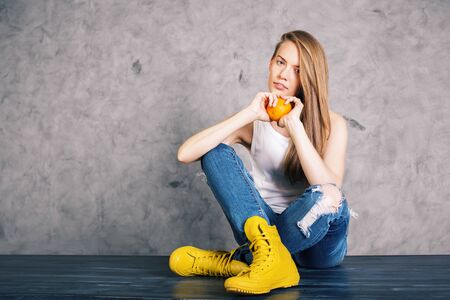 Pretty girl in yellow boots and with orange in hands sitting on wooden surface on concrete wall...