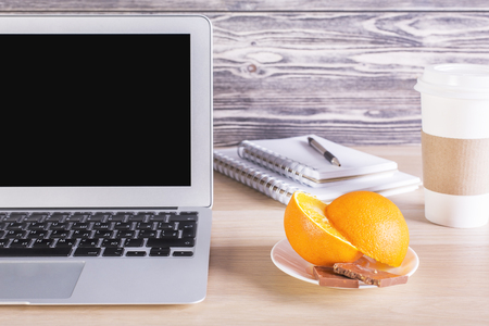 Closeup of desktop with blank laptop screen, orange slices and chocolate on saucer, coffee cup, notepads and pen. Mock upの写真素材