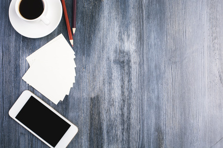 Top view of wooden desktop with blank smart phone, paper fan, coffee cup and pencils. Mock up