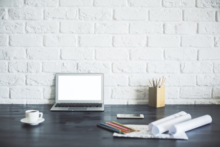 Blank laptop with white screen on wooden desktop with coffee cup, pencils, paperwork and other items on white brick background. Mock upの写真素材