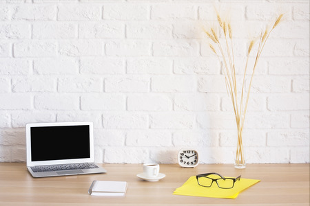 Bright workplace with blank laptop, notepad, coffee cup, clock, glasses on yellow paper sheet and wheat spikes on brick wall background. Mock upの写真素材