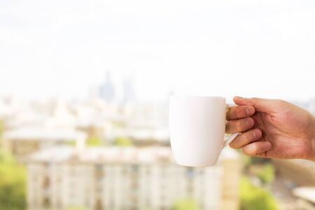 Hand holding blank white coffee mug on blurry city background. Mock upの写真素材