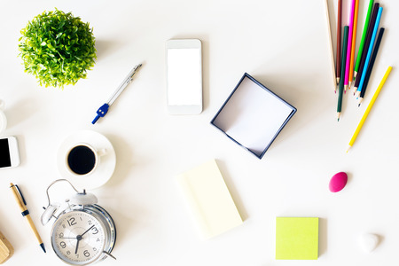 Top view of messy office desktop with blank white smart phone, plant, alarm clock and various stationery items. Mock upの写真素材