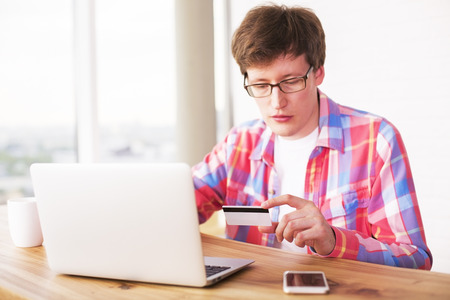 Online shopping concept with handsome european guy sitting at office desk looking at credit card and using laptopの写真素材