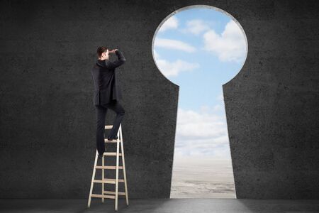 Success concept with businessman on ladder looking through keyhole shaped door with clear sky view in concrete roomの写真素材