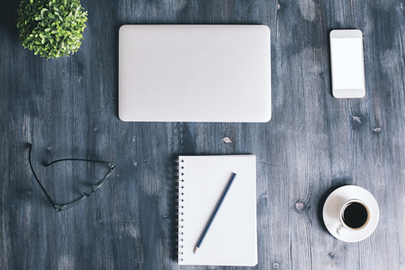 Top view of dark wooden office table with closed laptop, blank white smartphone, spiral notepad with pencil, glasses, coffee cup and plant. Mock upの写真素材