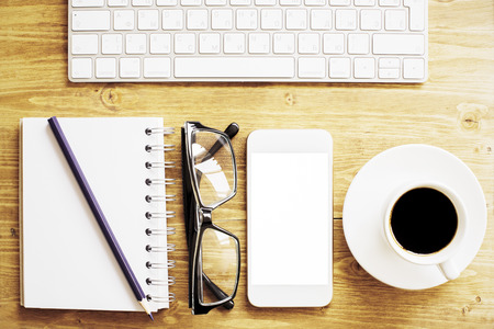 Top view of wooden office desktop with blank white smart phone, keyboard, spiral notepad with pencil, glasses and coffee cup. Mock upの写真素材
