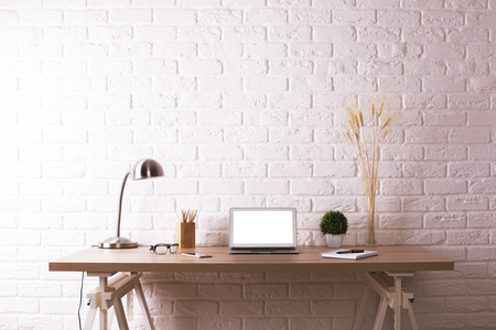 Front view of creative wooden designer desktop with blank white laptop, decorative plants, table lamp, glasses and stationery items on white brick wall background. Mock upの写真素材