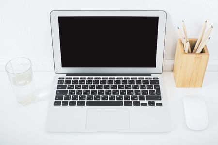 Front view of blank laptop on desktop with glass of water, pencils in box and computer mouse. Mock...