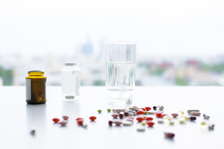 Closeup of white desktop with colorful pills, medicine bottles and glass of water on blurry city backgroundの写真素材