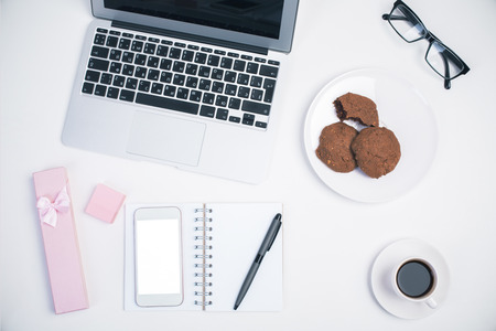 Top view of light girly office desktop with blank white smart phone, glasses, laptop keyboard, coffee cup, gift case, cookies on plate and other items. Mock upの写真素材