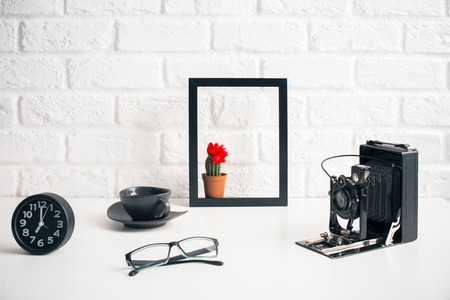 Creative white desktop with retro camera, cactus in frame, glasses, clock and coffee cup on brick wall backgroundの写真素材