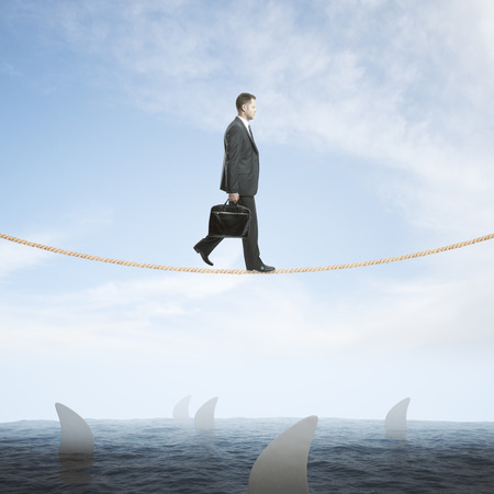 Risk concept with businessman walking on rope above sea full of sharks on sky backgroundの写真素材