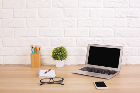 Front view of creative designer desktop with blank laptop, cell phone, glasses, plant and stationery items on white brick wall background. Mock upの写真素材
