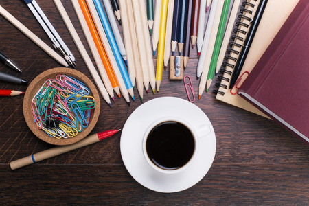Closeup of colorful pencils, pens, paper clips, notepad and book around white coffee cup on wooden...