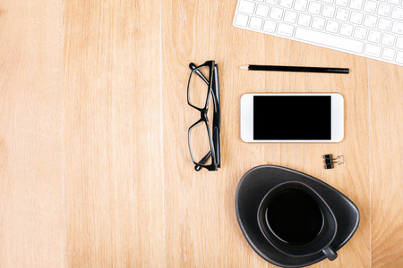 Top view of wooden office desktop with blank smartphone, coffee cup, spectacles, supplies and keyboard. Mock upの写真素材