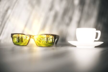 Blurry grey table with coffee cup and green forest reflected on sunglasses lens