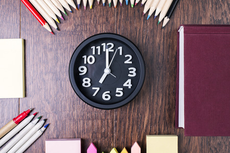 Top view of black clock, closed book, colorful pencils and other stationery items on dark wooden desktop. Close upの写真素材