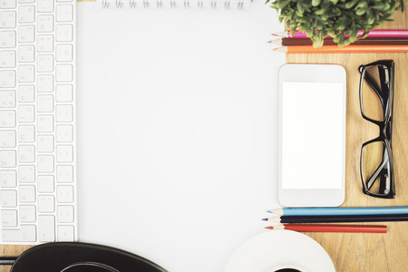 Closeup of wooden desktop with blank white smartphone, paper sheet, coffee cups, glasses, colorful pencils, keyboard and plant. Mock upの写真素材