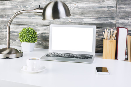 Closeup of creative designer workplace with blank white laptop screen, smartphone, coffee cup, table lamp, plant, books and pencils on wooden wall background. Mock upの写真素材