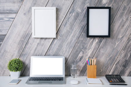 Creative designer workplace with blank white laptop computer, plant, smartphone, water glass, calculator, stationery items and two picture frames hanging above on wooden background. Mock upの写真素材