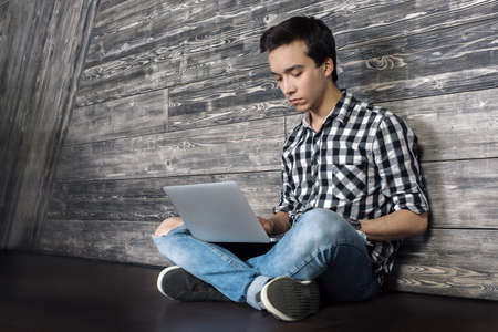 Stylish young man with laptop sitting on floor in wooden interiorの写真素材