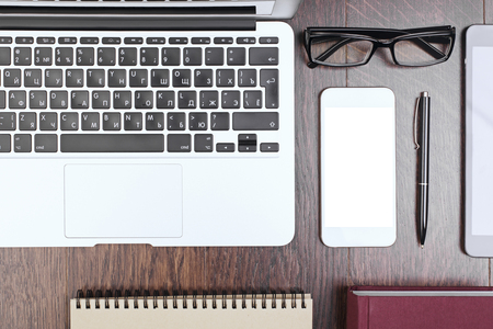 Top view and closeup of wooden office desktop with laptop keyboard, blank white mobile phone screen, spiral notepad, book, pen and glasses. Mock upの写真素材