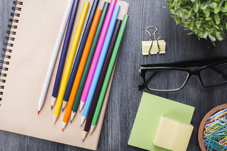 Close up of colorful pencils, spiral notepad, glasses, stickers, peg, paper clips and decorative plant on wooden desktop. Top viewの写真素材