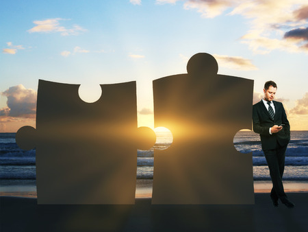 Businessman leaning on abstract giant puzzle pieces and using smartphone at the beach with setting sunの写真素材