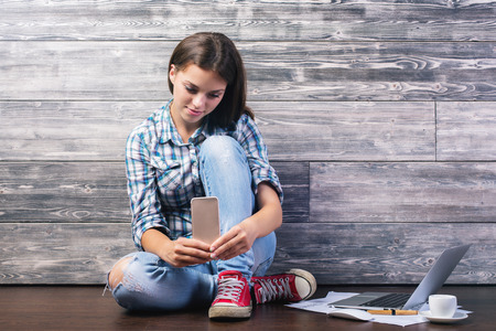 Casual young woman sitting on wooden floor next to laptop, paperwork and coffee cup, using mobile phoneの写真素材