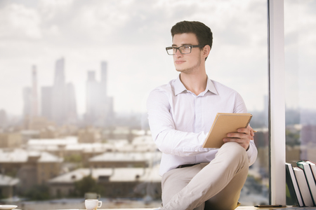 Portrait of handsome young businessman with spiral notepad in hand sitting on windowsill with coffee cup and books. Blurry city view in the backgroundの写真素材