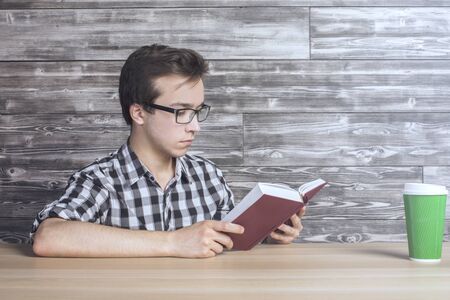 Smart young guy in glasses reading reading book at wooden table with green take-away coffee cup. Textured wooden wall in the backgroundの写真素材