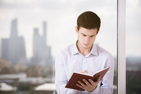 Portrait of handsome young businessman at workplace writing in hardcover notepad against window with blurry city viewの写真素材