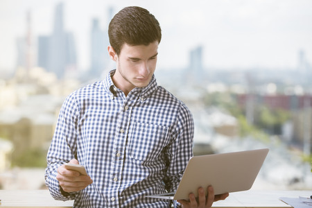 Portrait of handsome young businessman with cellphone in hand looking at laptop screen on blurry city backgroundの写真素材