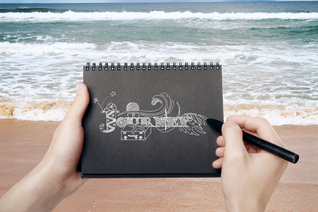 Male hands holding black spiral notepad and drawing journey sketch on seaside background. Traveling conceptの写真素材