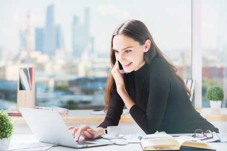 Portrait of smiling happy business woman at office desk talking on cellular phone and using laptop computer. Blurry city view in the backgroundの写真素材