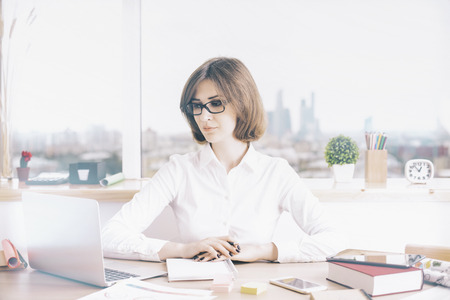 Attractive businesswoman sitting at office desktop with laptop and other items. Window with city view in the backgroundの写真素材