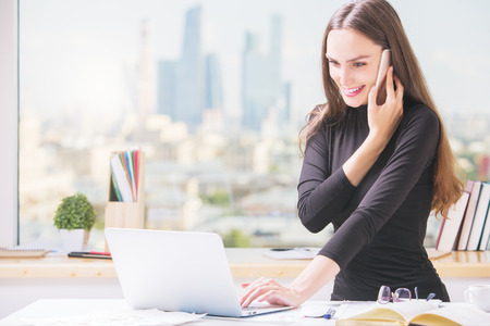 Portrait of smiling young businesswoman at office desk talking on cellular phone and using laptop computer. Blurry city view in the backgroundの写真素材
