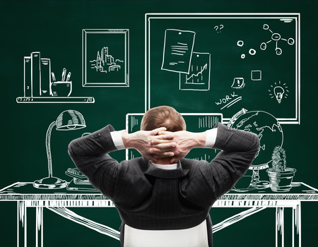 Back view of young businessman in suit relaxing in creative drawn office on chalkboard backgroundの写真素材
