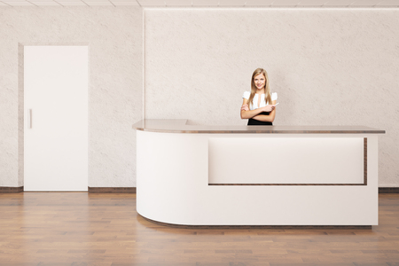 Smiling european businesswoman standing at reception desk in interior with wooden floor, concrete walls and white door. 3D Renderingの写真素材
