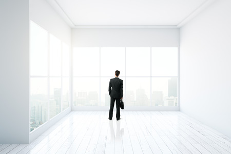 Thoughtful businessman standing in minimalistic interior design with white walls, floor, ceiling and panoramic windows with city view. 3D Renderingの写真素材