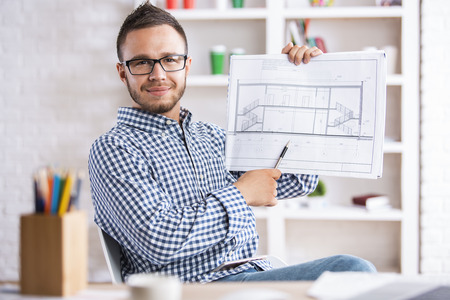 Smiling young engineer pointing at construction project with pen while sitting at modern bright...