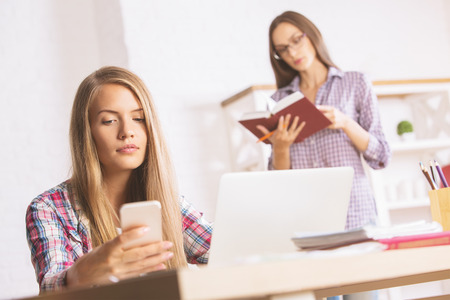 Two pretty young ladies at workplace using cellular phone and reading bookの写真素材
