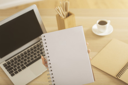 Closeup of female hands holding blank spiral notepad above workplace with laptop computer, coffee cup and supplies. Mock upの写真素材